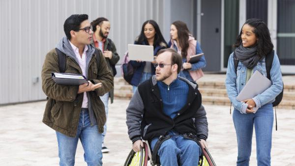 A multiracial group of young adults, university students, hanging out outside a campus building, conversing. The friends include a man with cerebral palsy in a wheelchair. Three of the students are walking and conversing in the foreground while three others are out of focus in the background.